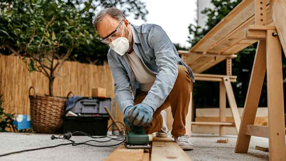 Man wearing a mask and safety goggles is sanding wooden furniture 