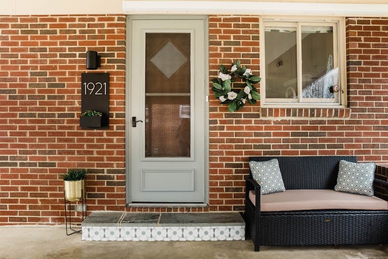 full entry way featuring gold plant stand, tiled step, patio bench, house number plant box and sconce light, and wreath