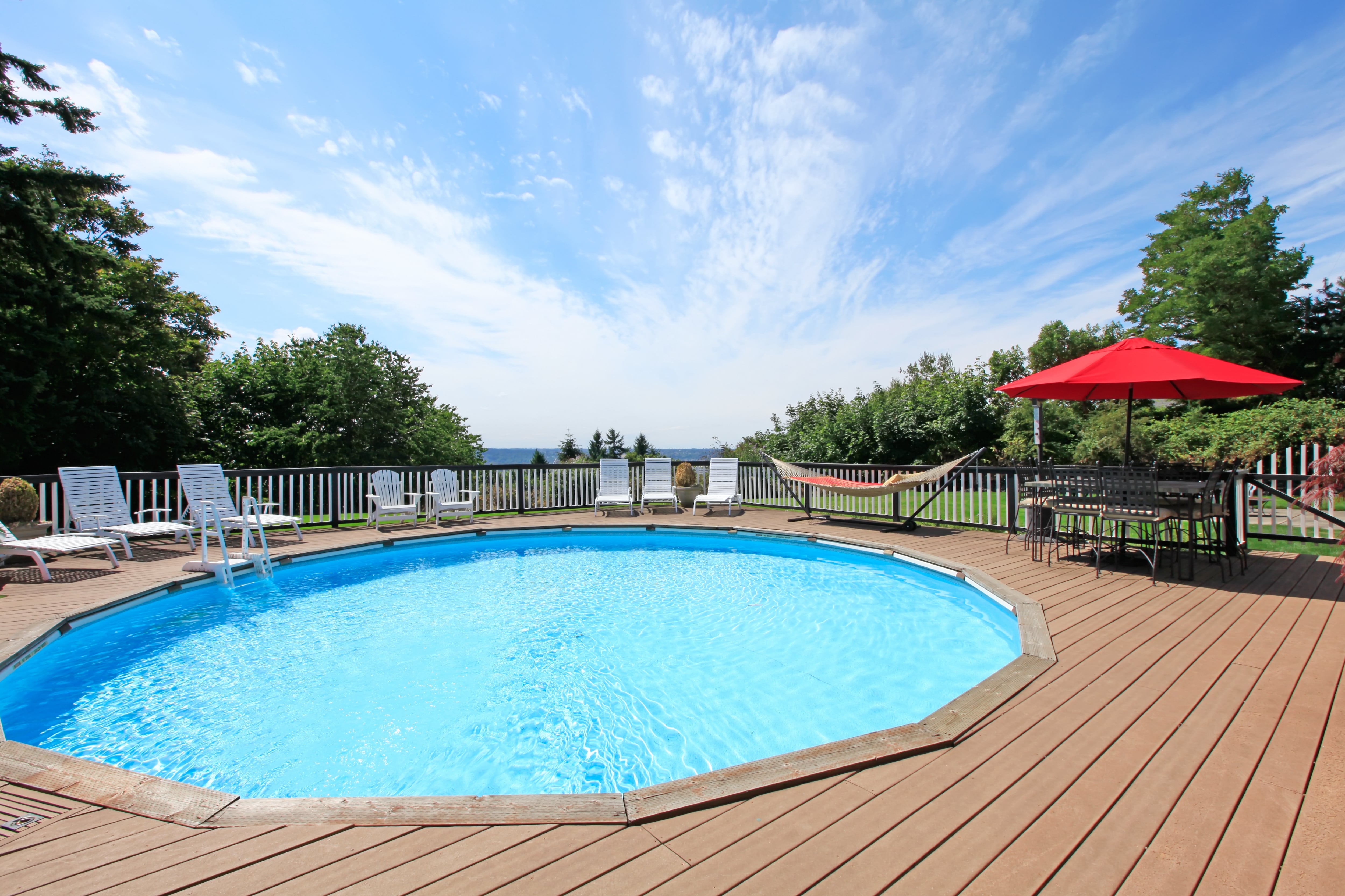 An above ground pool and surrounding deck with lounge chairs, hammock, and patio table 