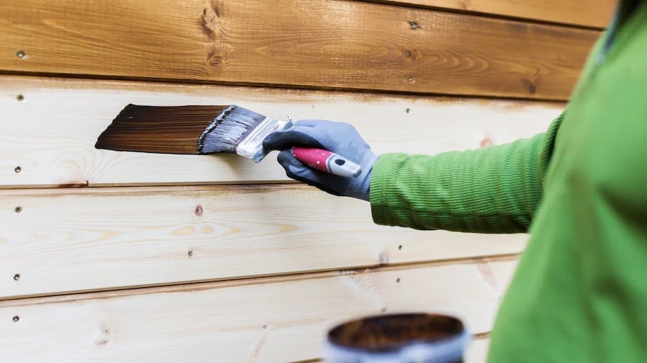 A painter wearing a gloves staining a wooden wall with a paintbrush