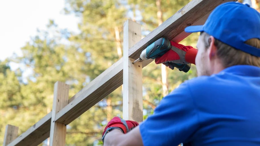 A man in a blue shirt and hat drilling screws to build a wall frame