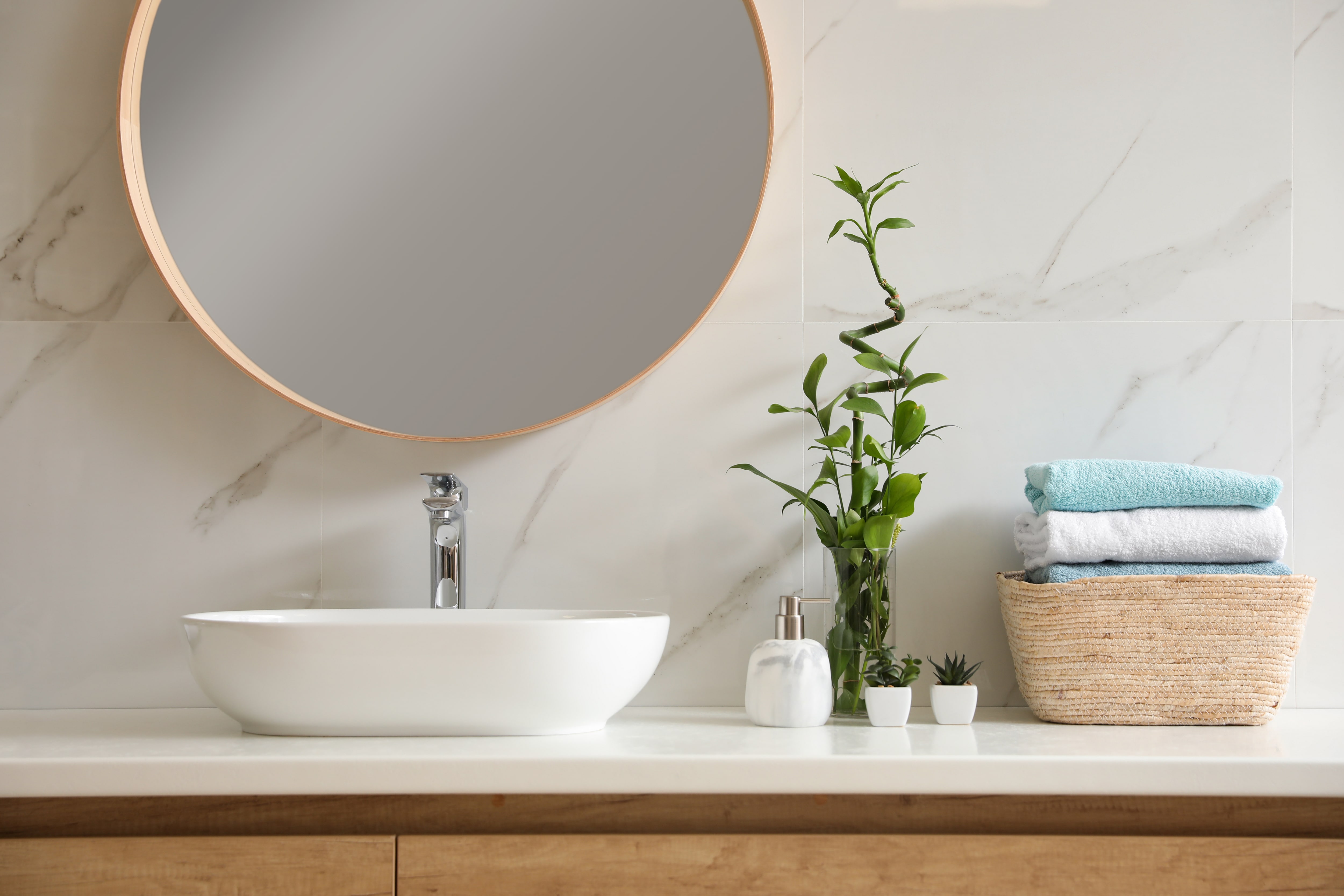 Modern bathroom with marble slab backsplash, round mirror, plants, vessel sink, and a woven basket with towels