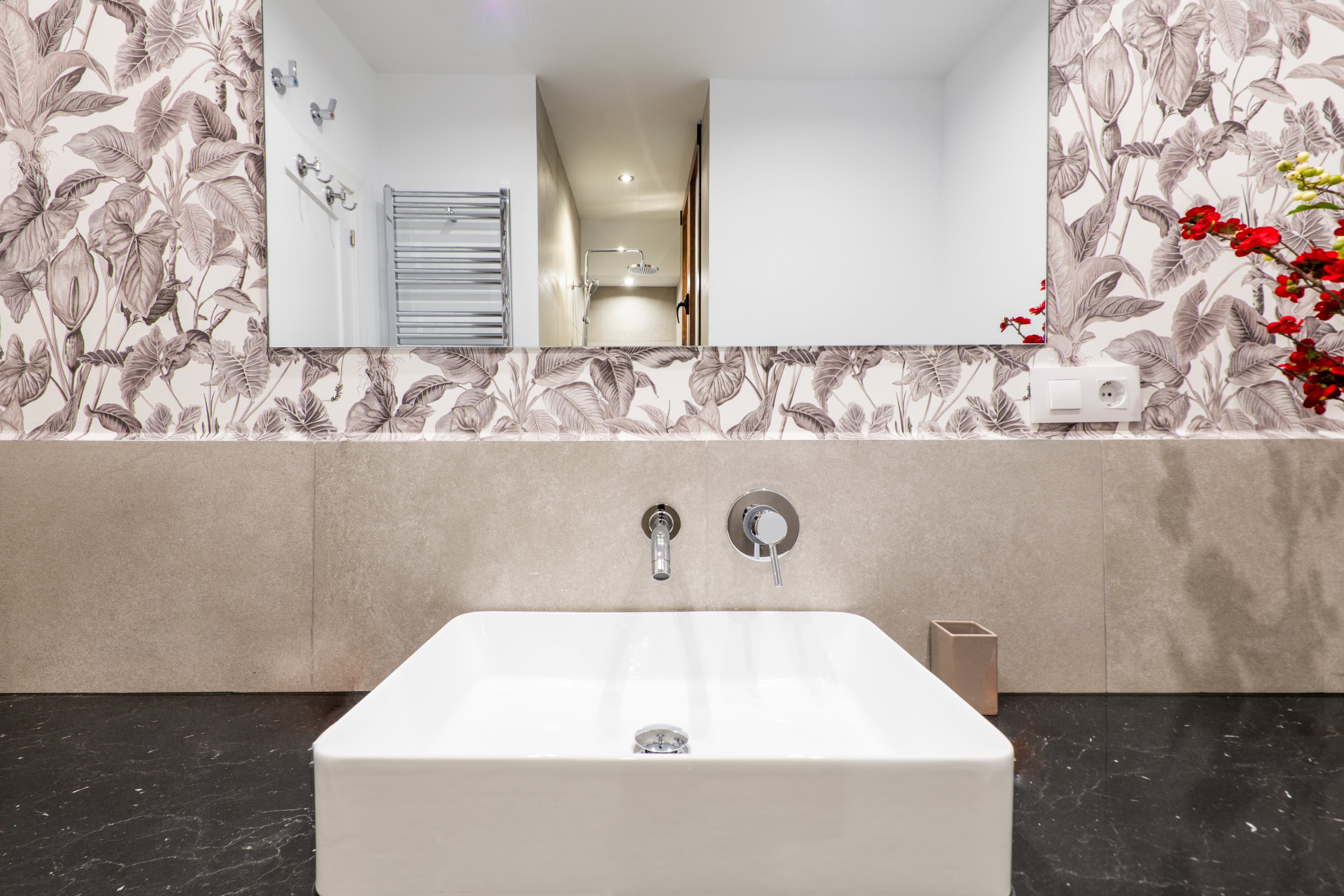 Modern bathroom with black countertops, white sink, chrome fixtures, square mirror, and wallpaper backsplash