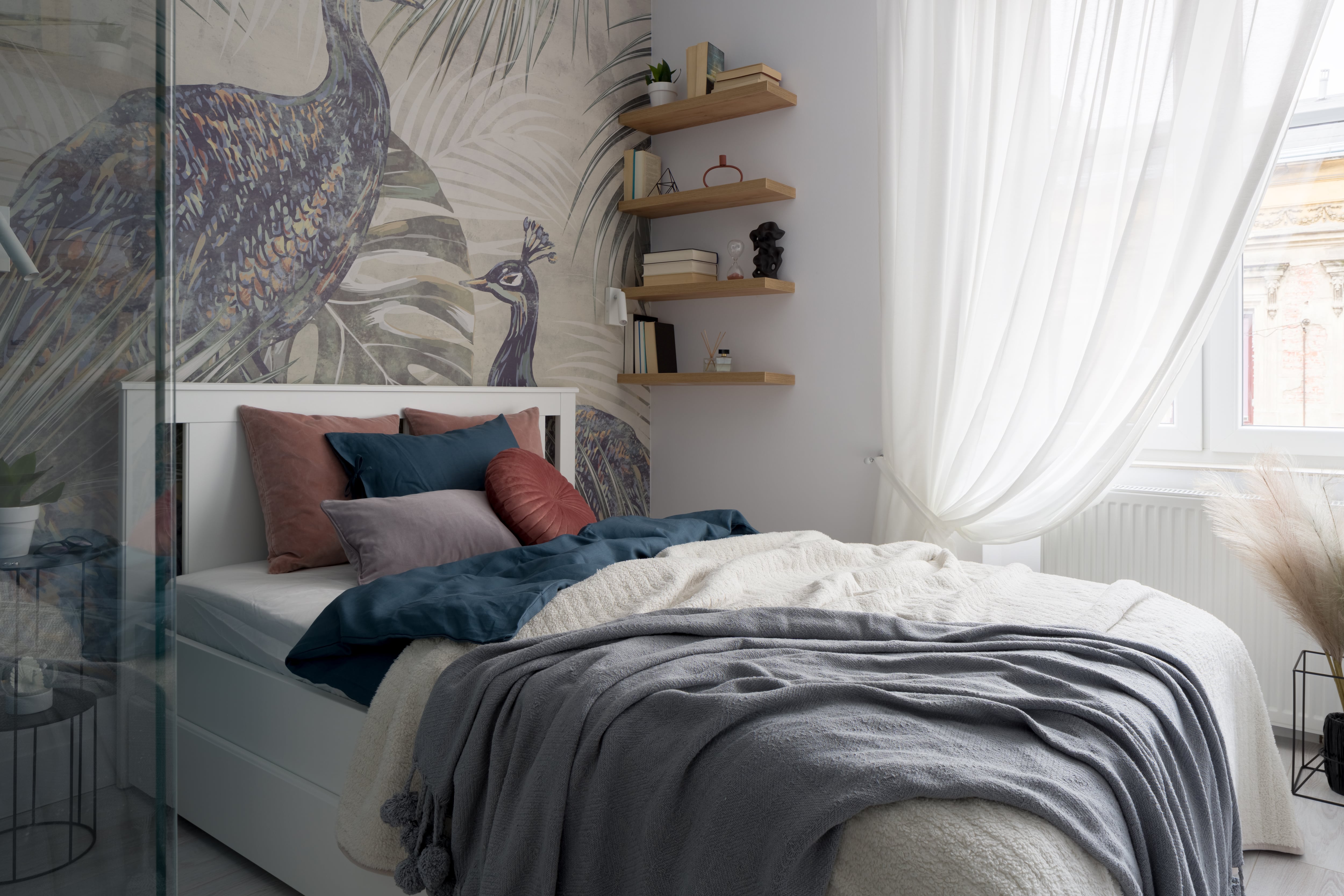 Small bedroom with a window, cozy bed, floating shelves, and peacock-patterned wallpaper