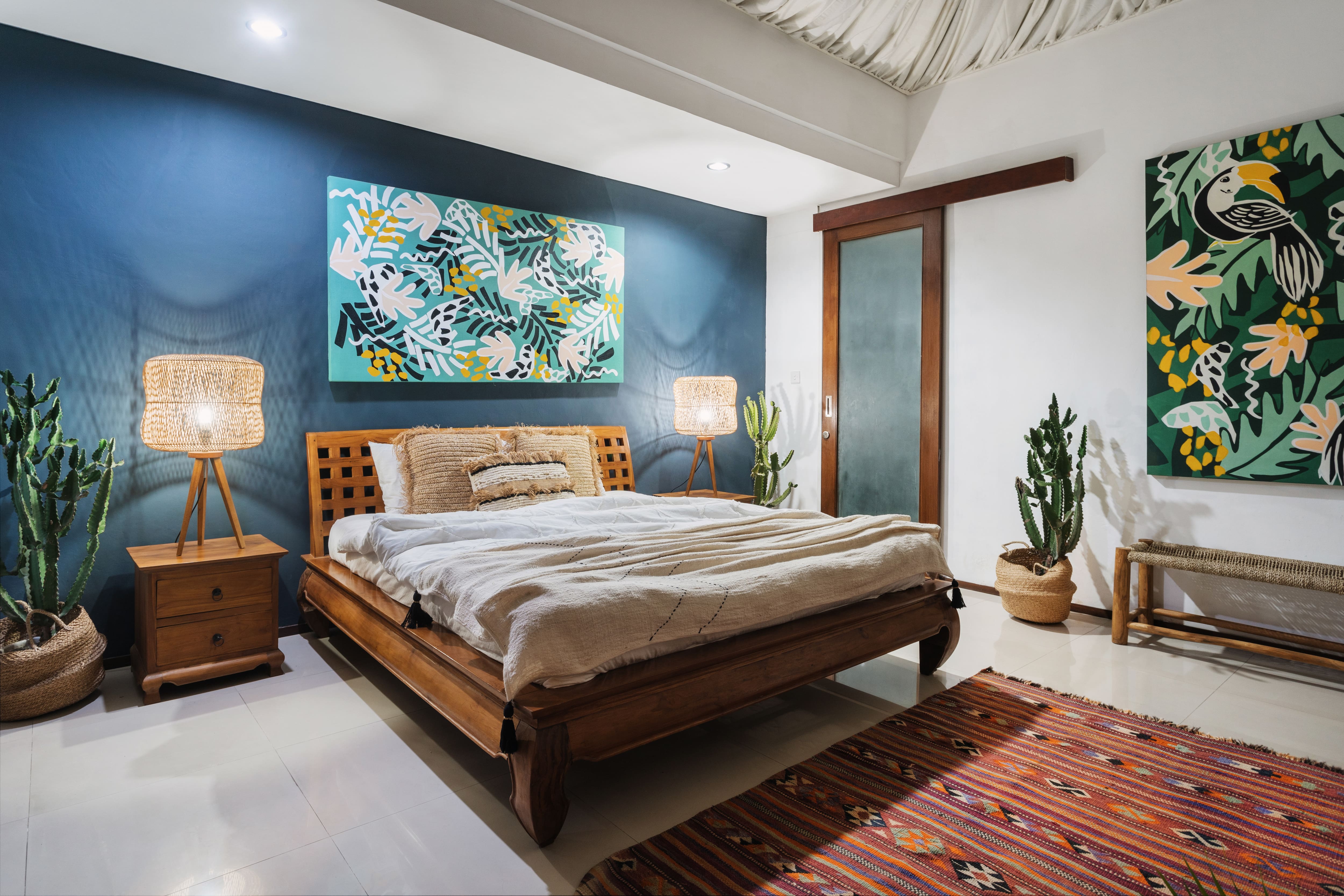 Eclectic bedroom with blue accent wall, large cactus, rattan lamps, wooden bed and nightstands, and oversized tropical art