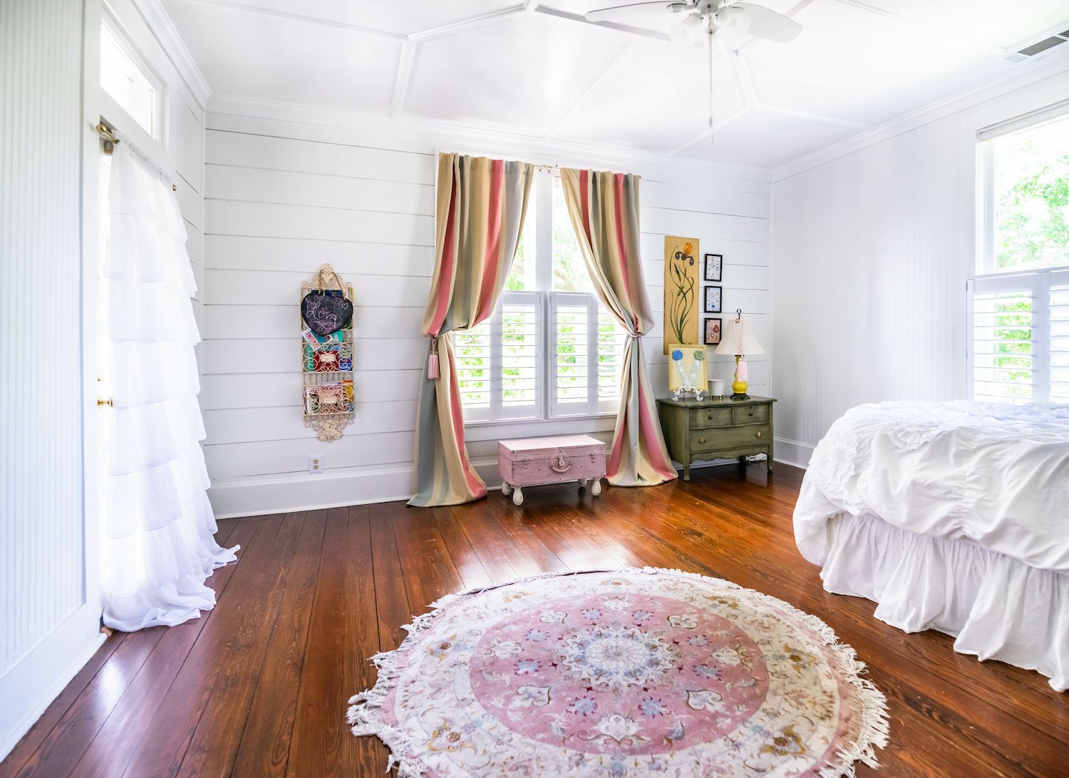 Bedroom with white shiplap walls, hardwood floors, tassel rug, and ruffled white and colorful striped curtains