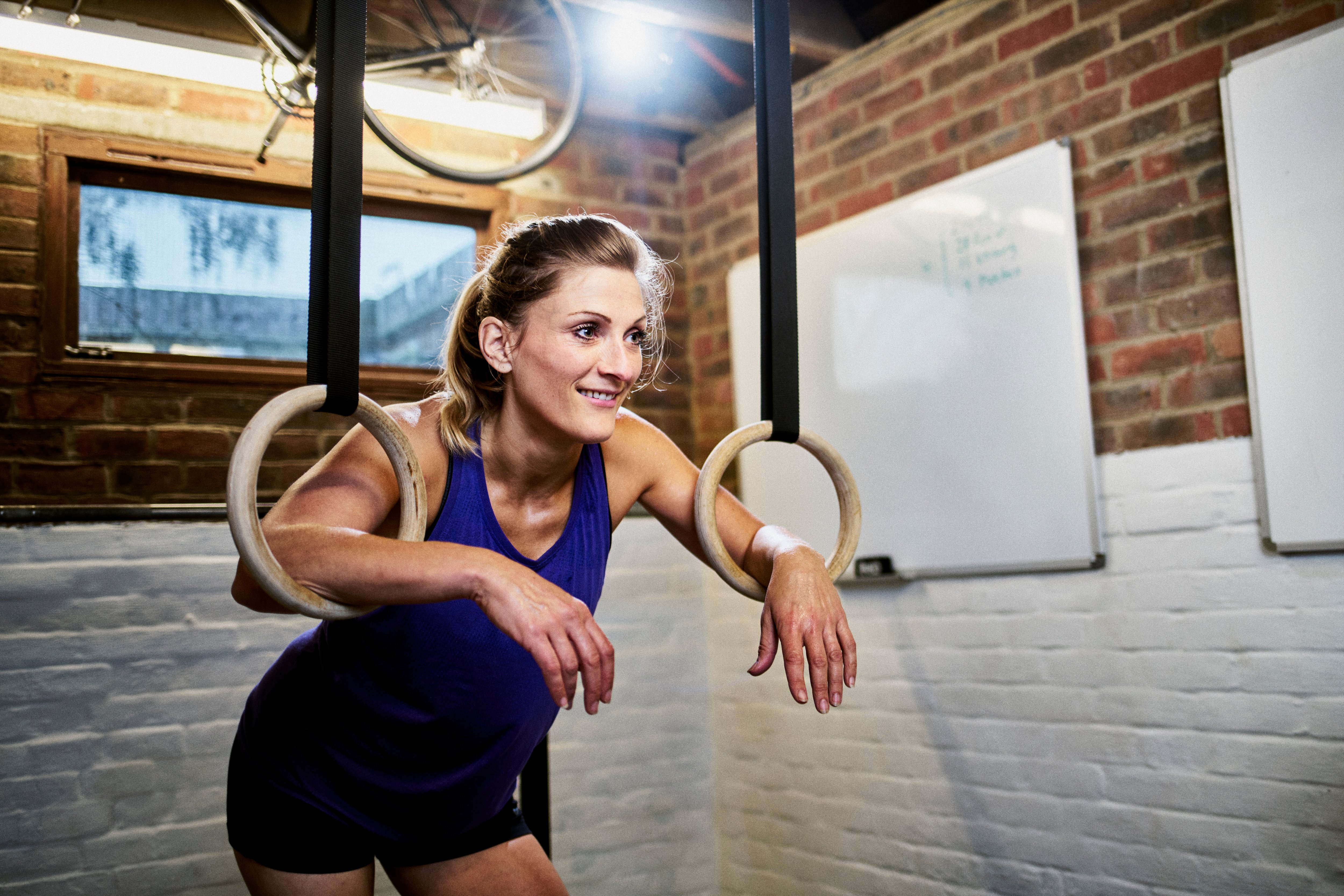 Woman in gym clothes with her arms through gymnastic rings in garage home gym and lights hanging in the background