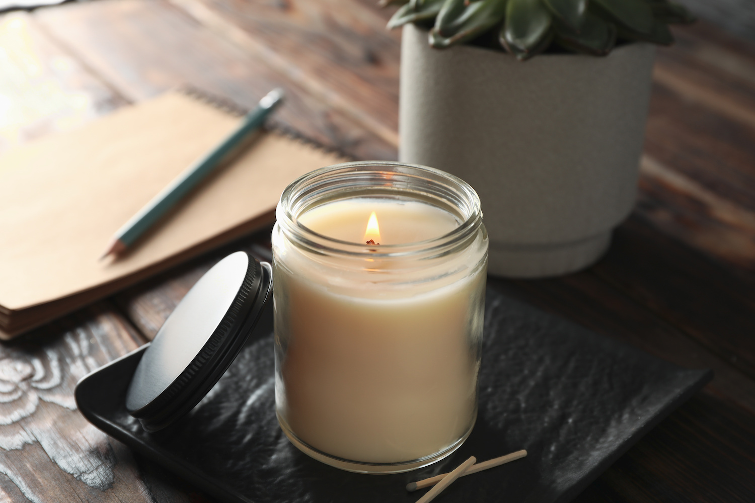 Dark wooden desk with a large white candle that’s lit in a glass jar next to a notebook and succulent