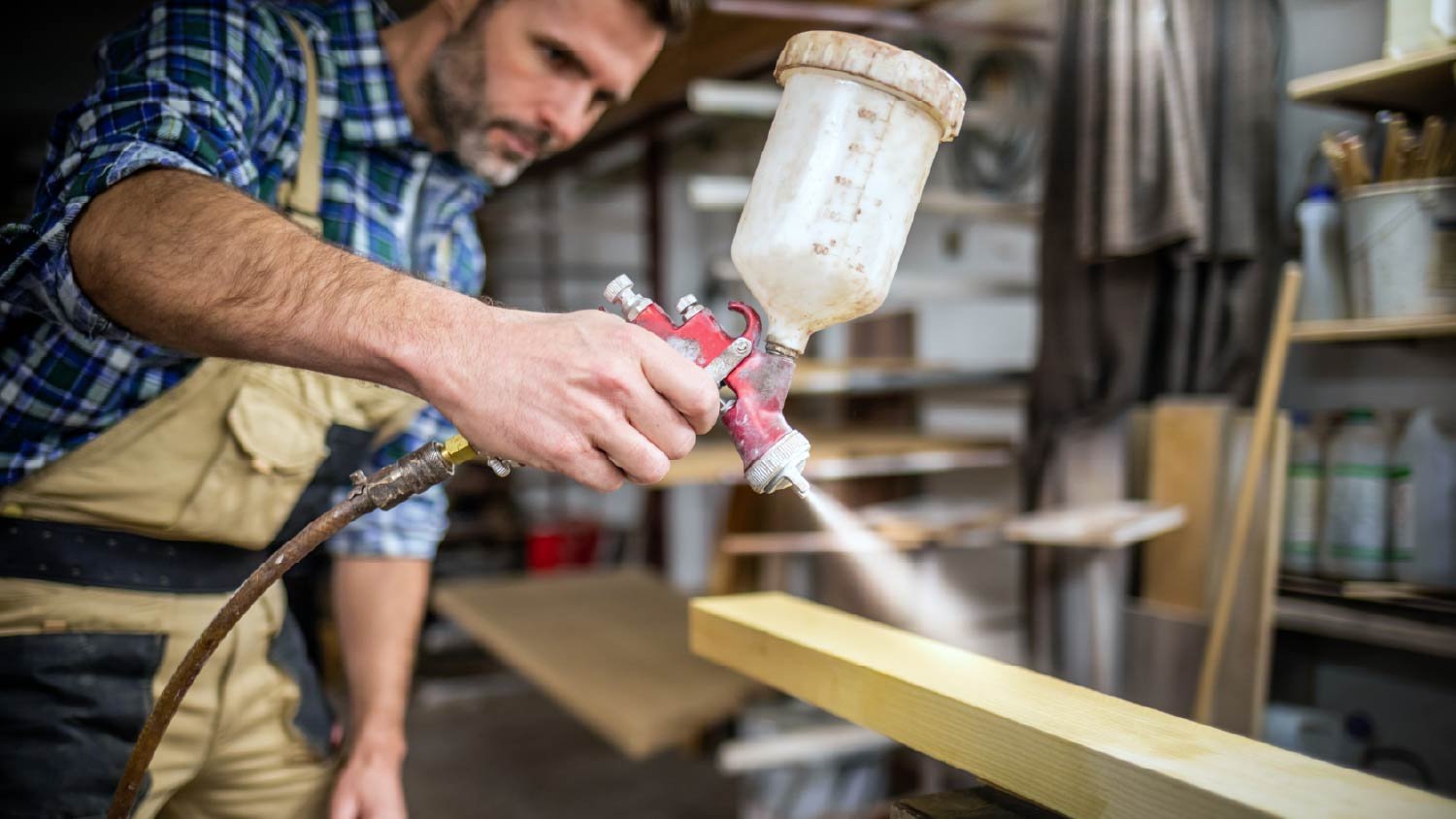 Carpenter with paint spray gun painting wooden plank