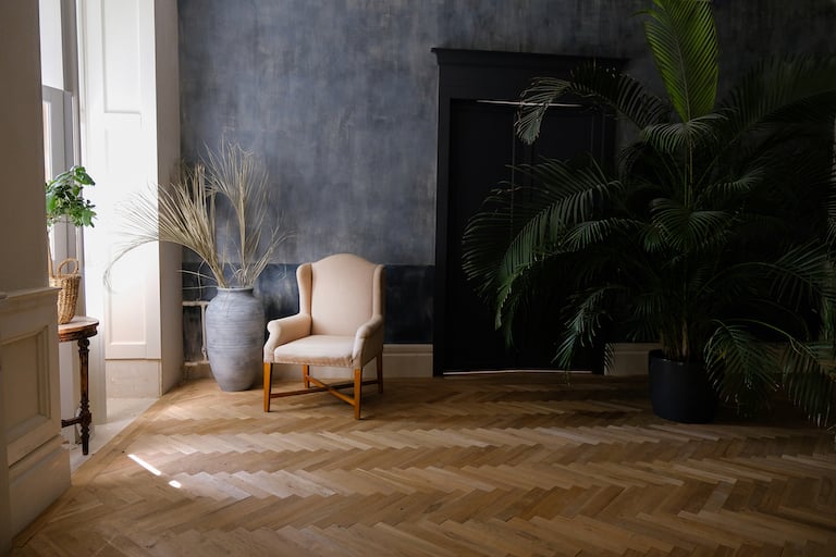 off-white accent chair sits in corner by window in room with dark blue walls and diagonal plank wood floors