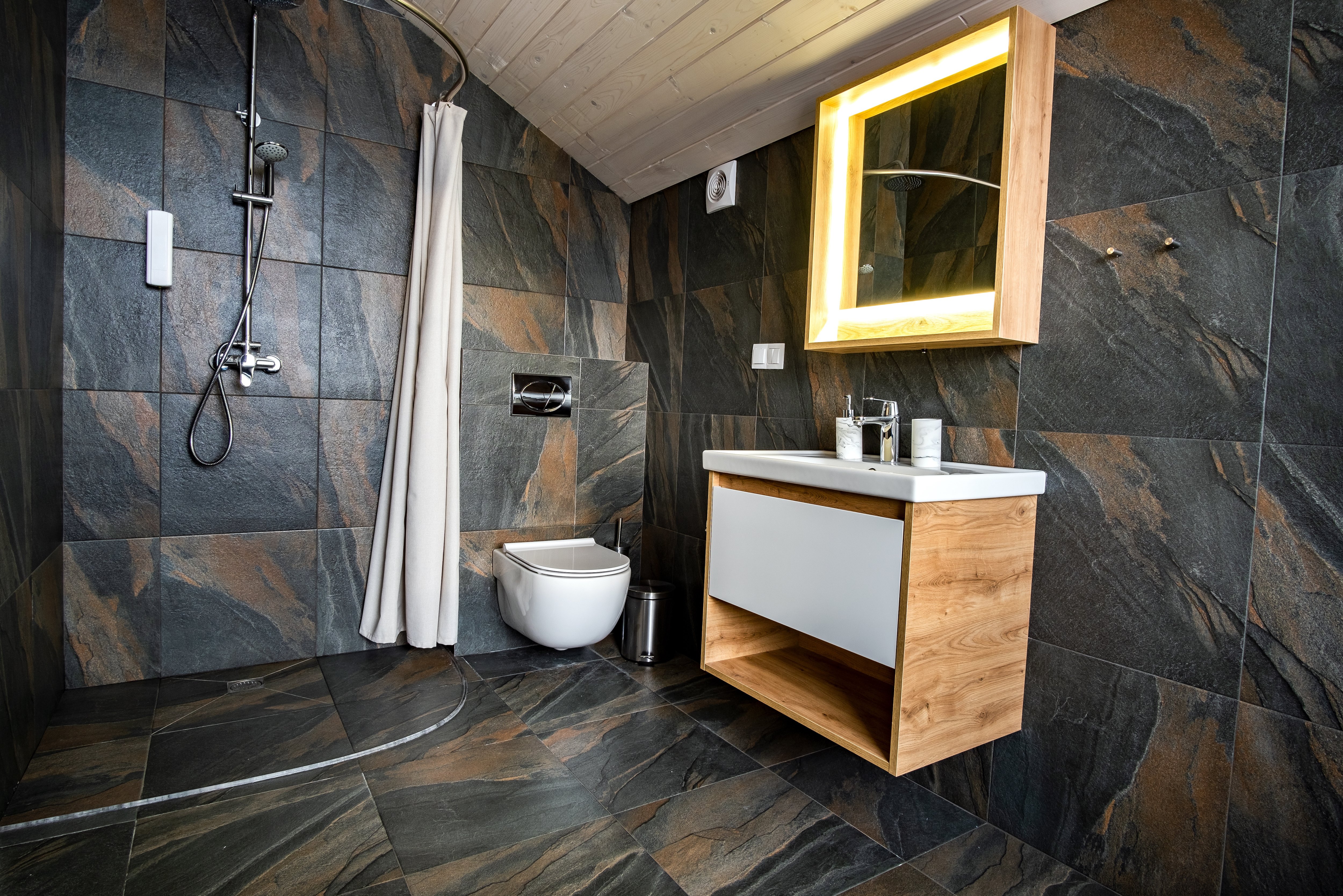 Bathroom with a doorless shower, wall-hung toilet, wood vanity and mirror, and black-and-brown tiles on the floor and walls