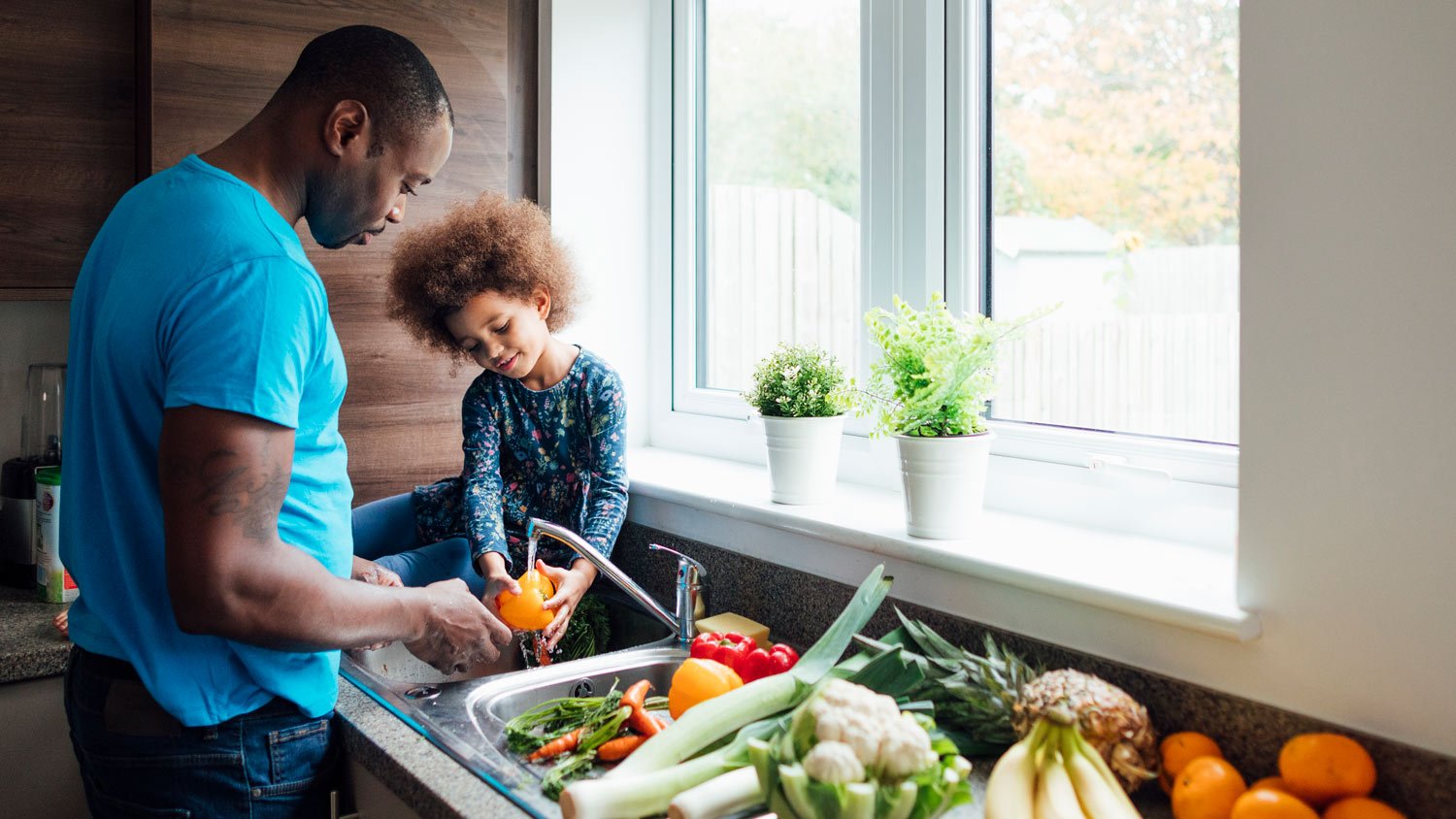 Wide shot of a father and his little girl washing fresh vegetables in the sink at home