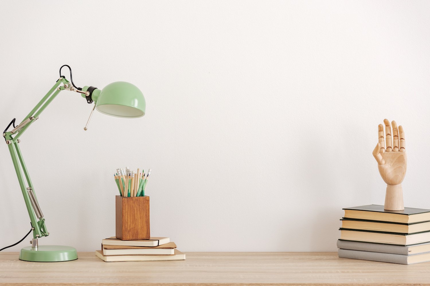 White wall with a wooden desk, mint green desk lamp, wooden pencil holder with stacks of books