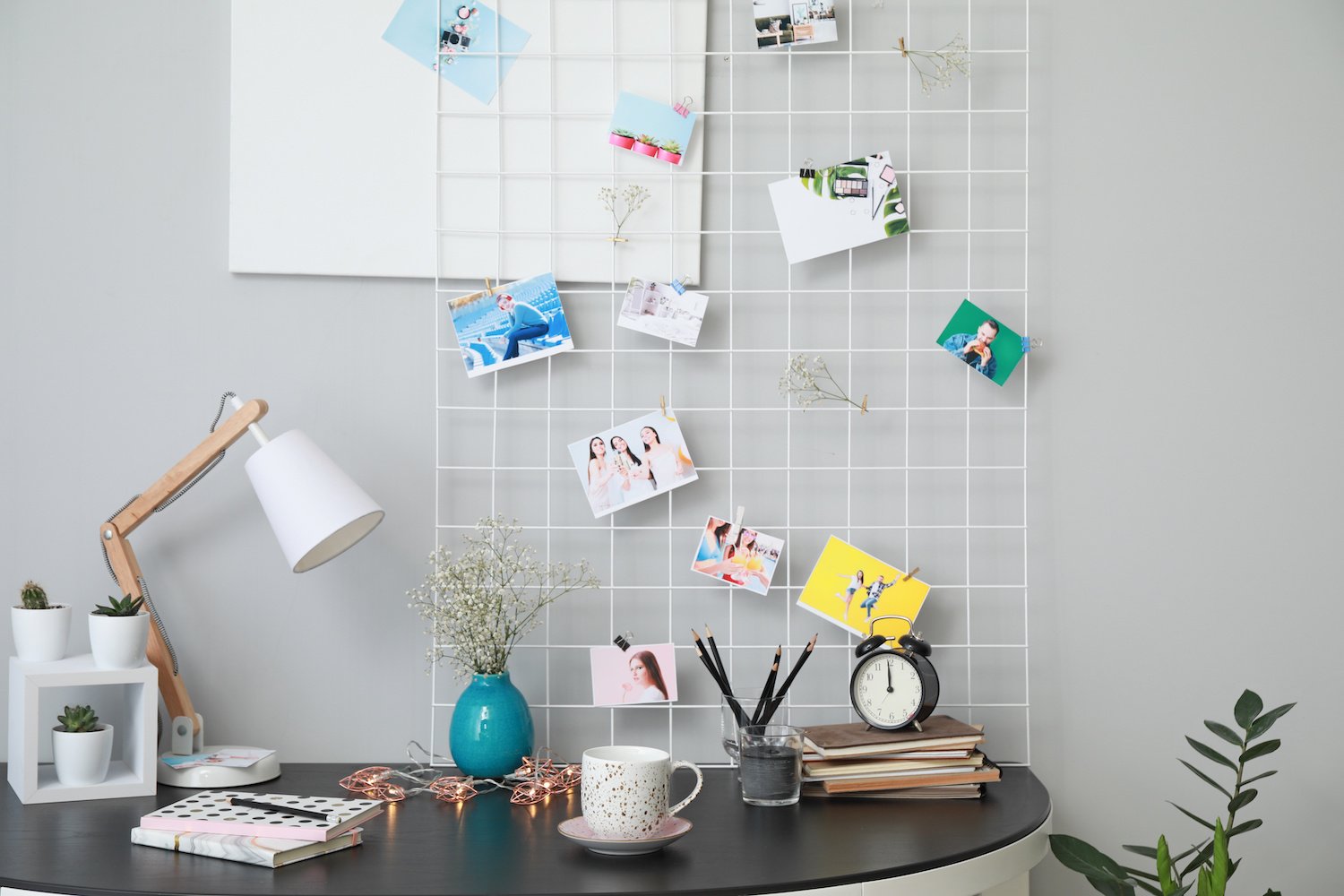 Black desktop with a white grid standing against a gray wall with pictures hanging from it next to books, a plant in a vase and desk lamp