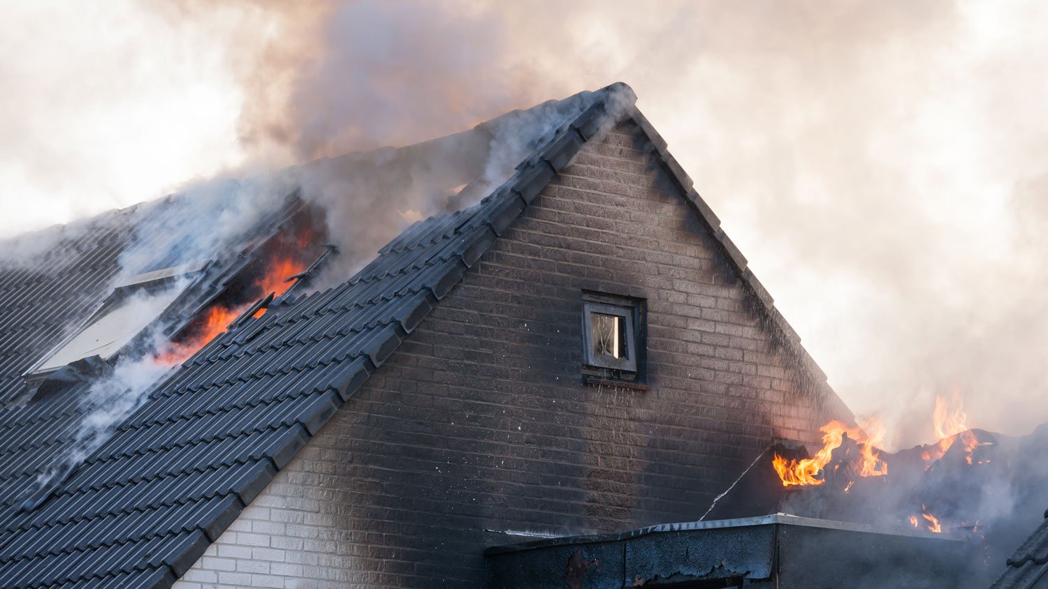 Fragment of a sooty white brick home that is on fire with flames and smoke