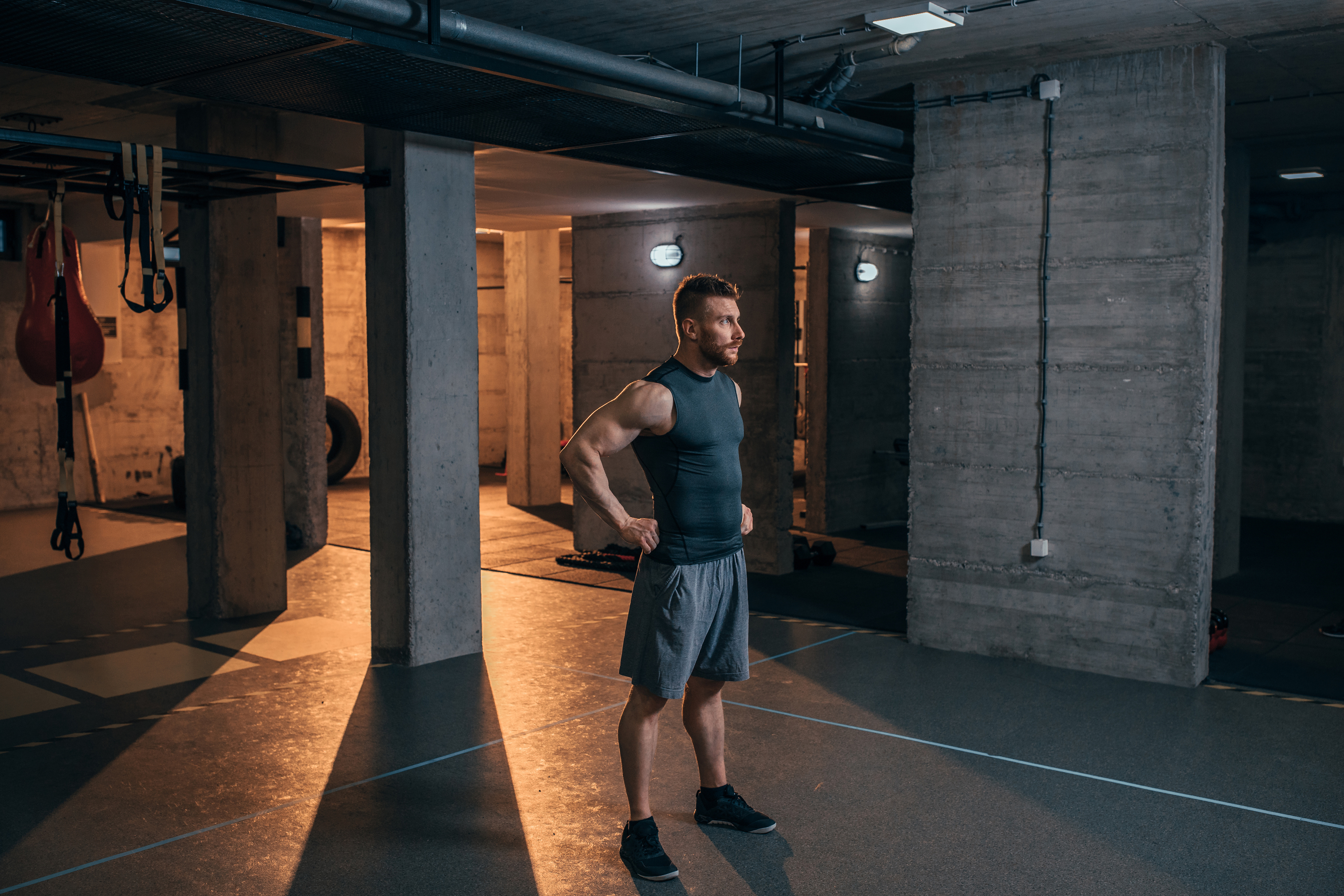 Man in gym clothes stands with his hands on his hips in an industrial basement home gym with concrete walls and weight machines