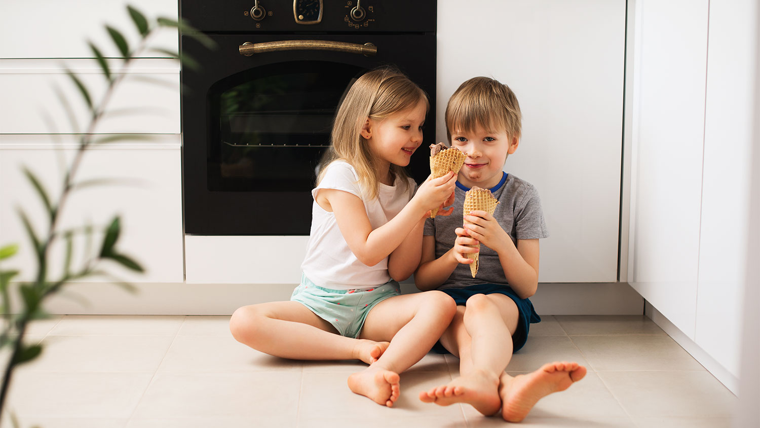 Two kids sitting on kitchen floor eat ice cream