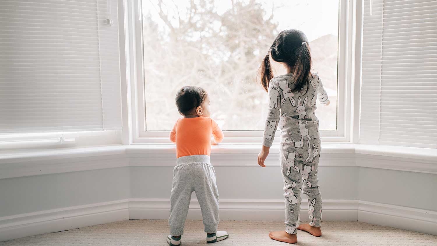 Siblings wearing pajamas are looking out the room window