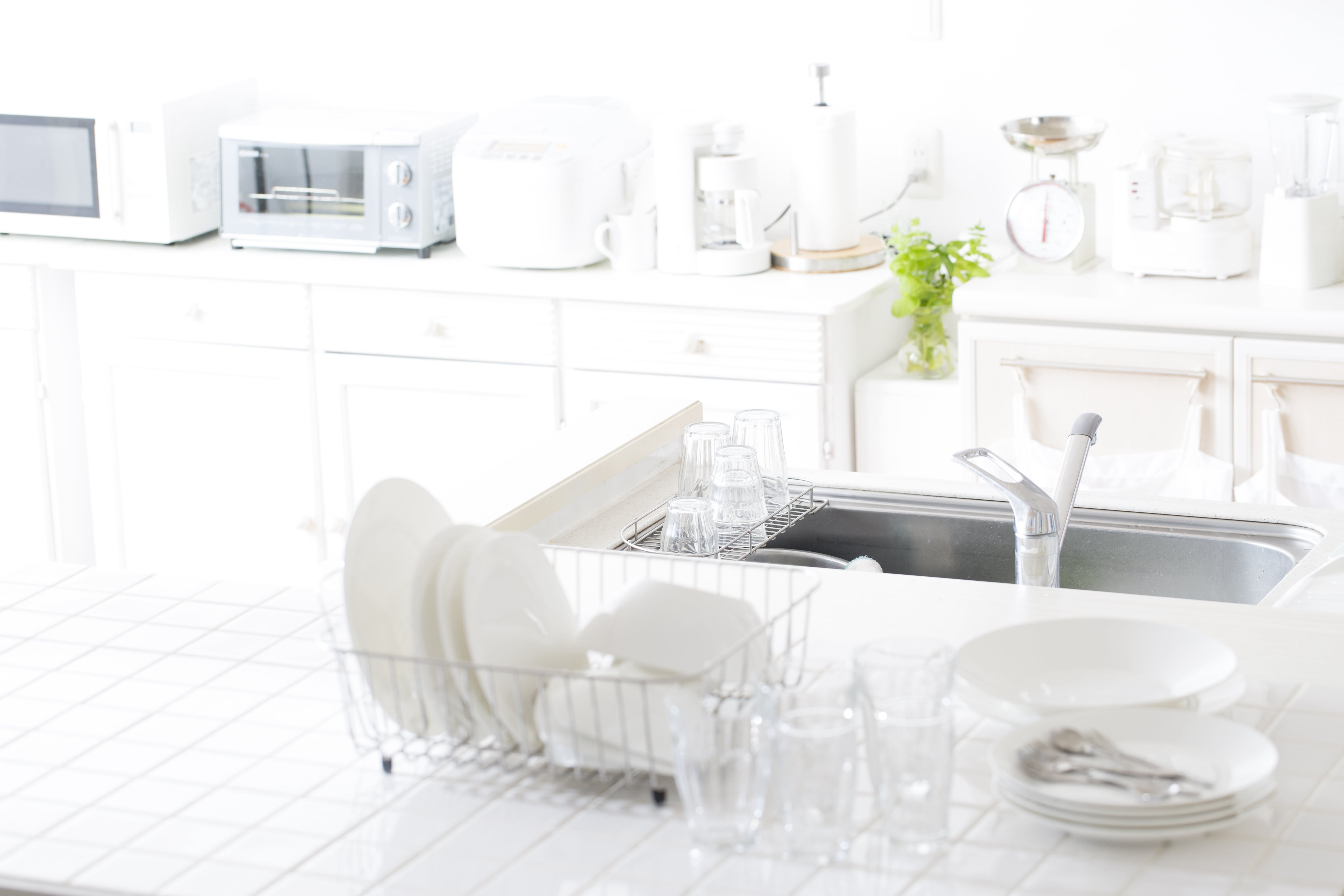 All-white kitchen with white tile countertops and various white dishes piled around a stainless sink 