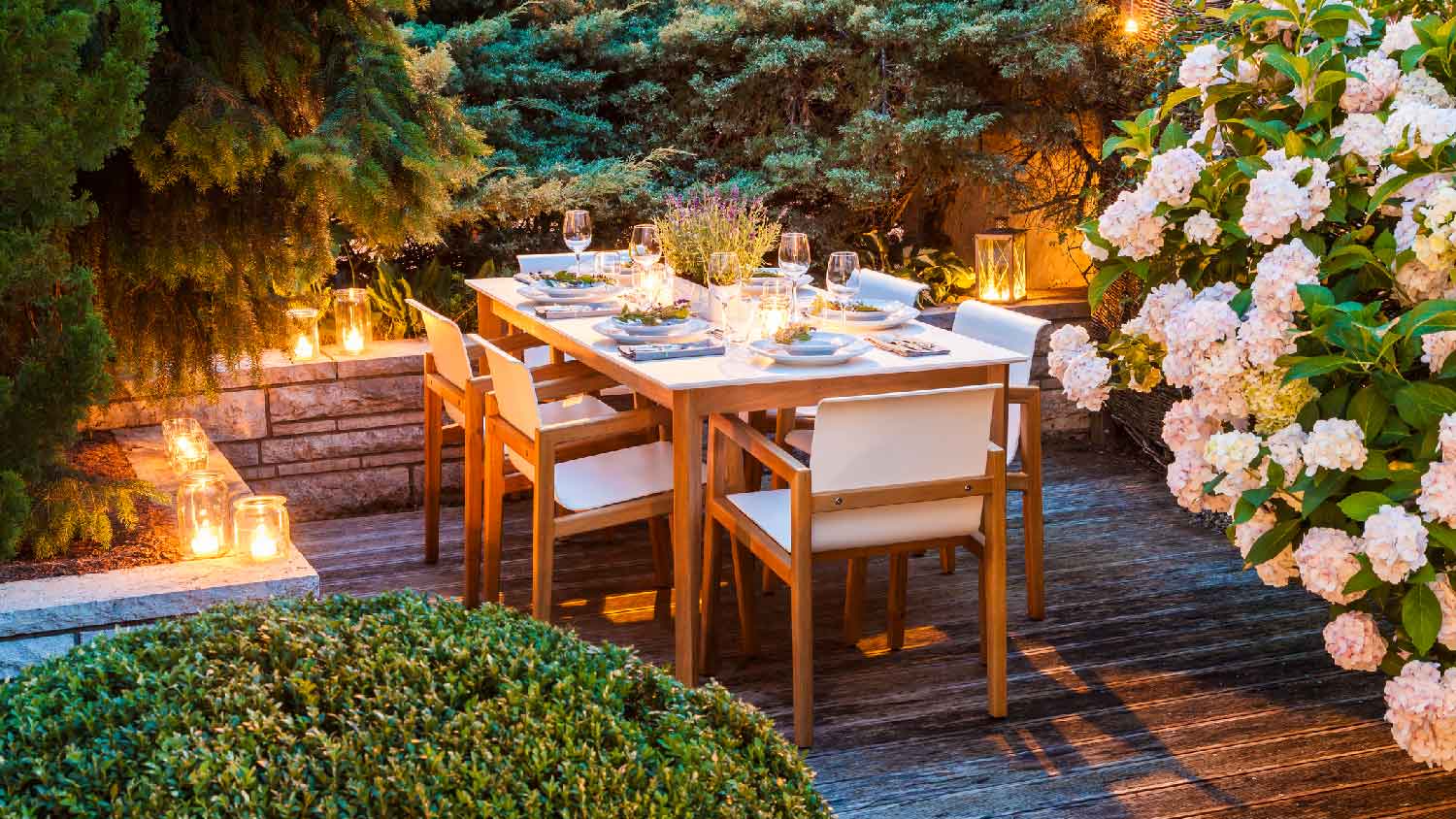 A laid table on a lighted terrace surrounded by trees and flowers