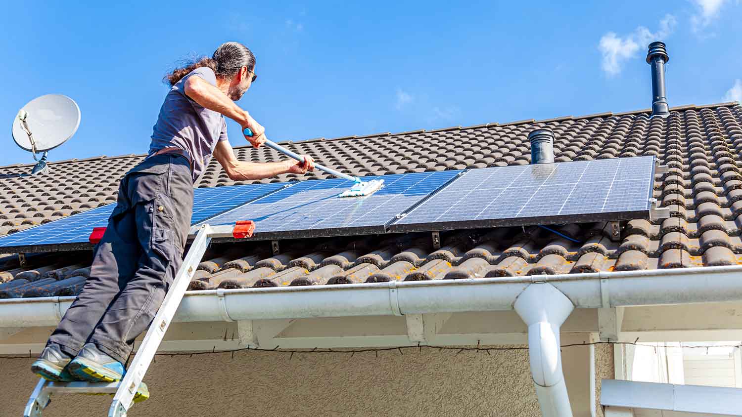 Man on a ladder using a long-handled brush to clean solar panels