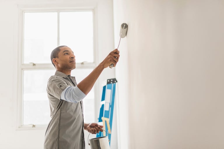 side shot of man painting white wall with white paint, holding roller brush and paint can with blue ladder in background