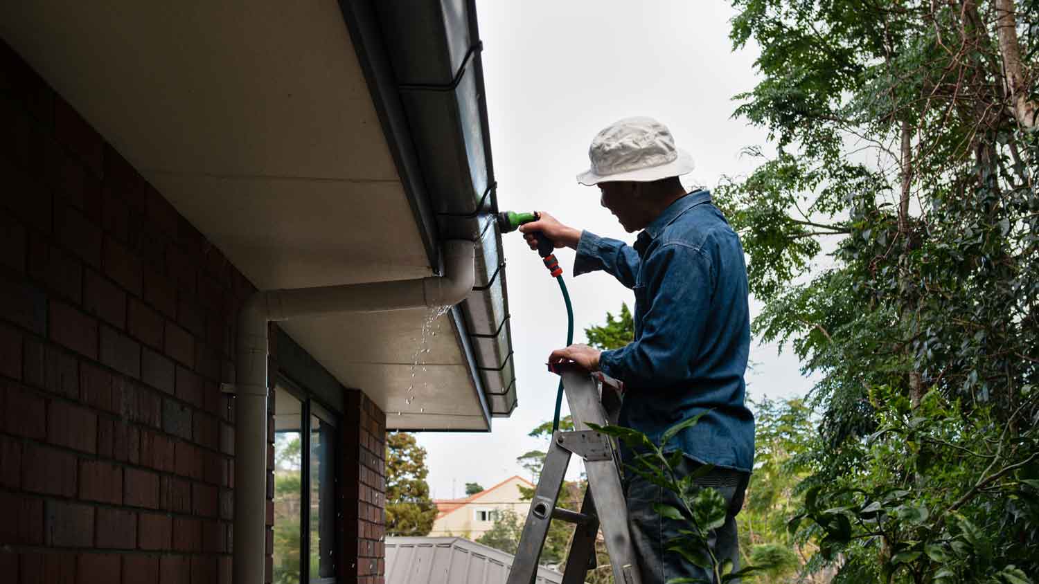 A man on a ladder rinsing gutters with a water hoses