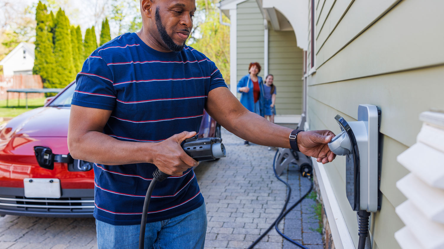 African-American father holding EV charger, ready to plug it into red car outside their home