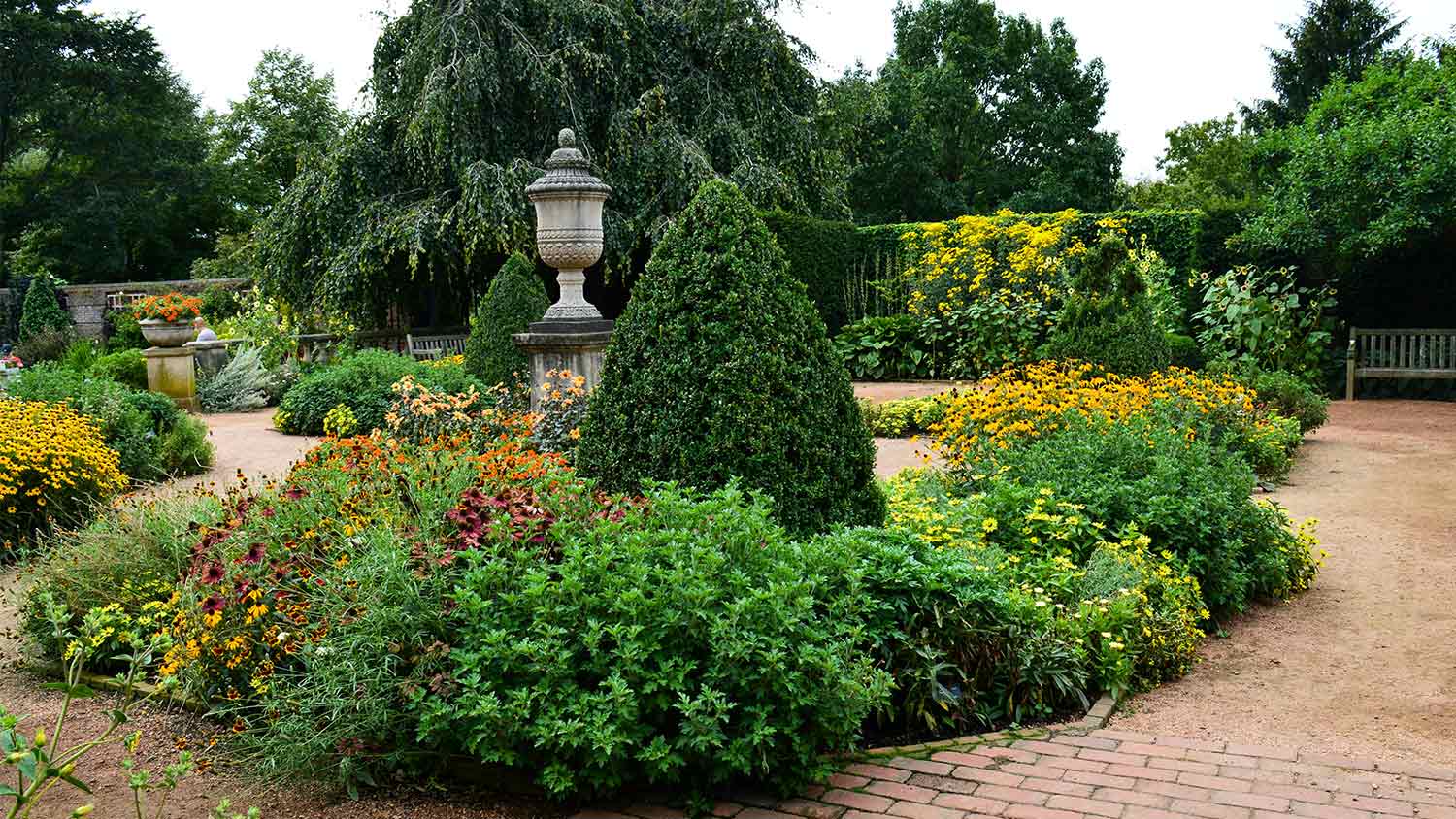 A garden with a mixture of flowers, shrubs, trees, statues, and brick walkways