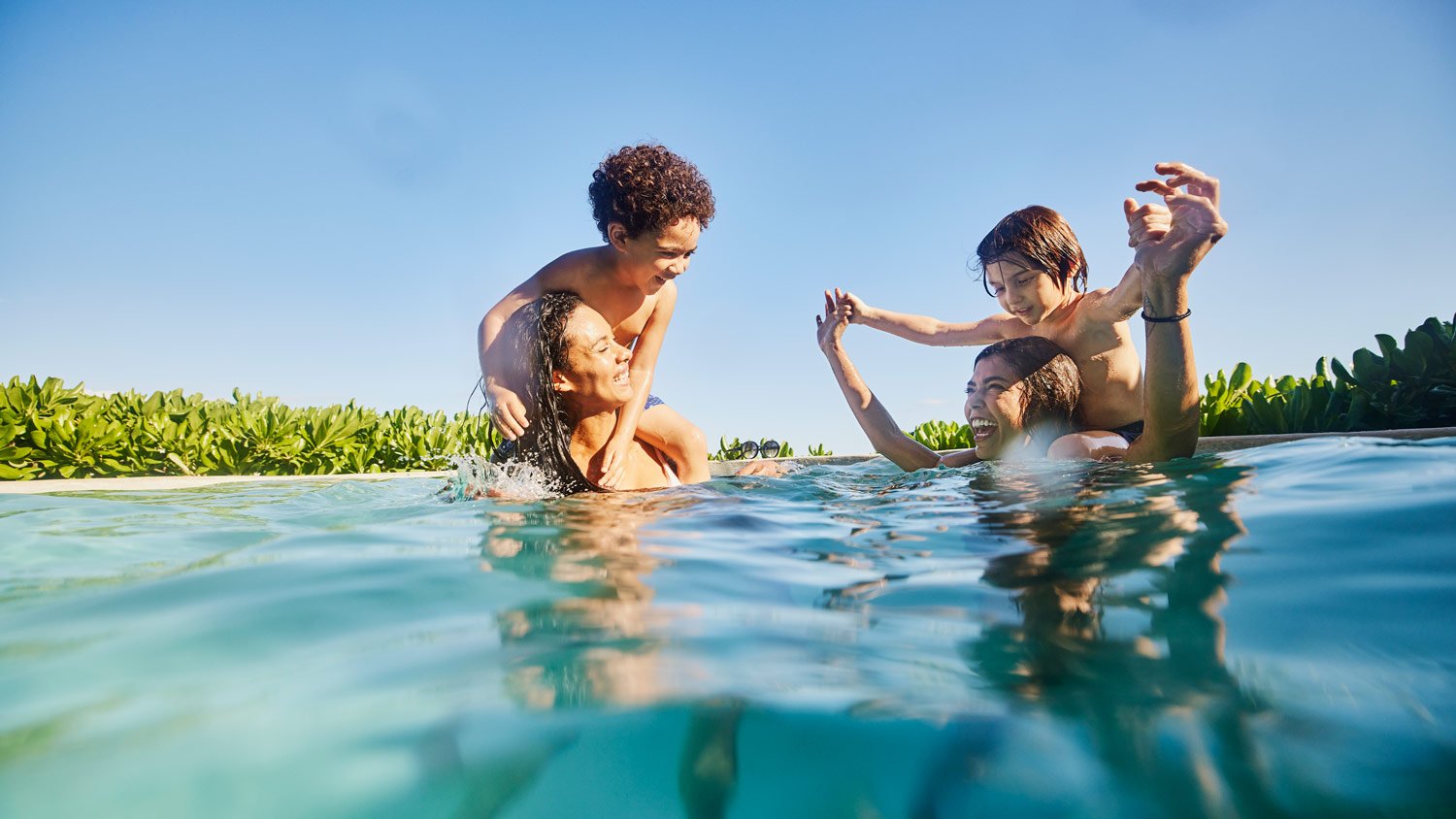 Wide shot of smiling sons riding on mothers backs while playing in pool at tropical resort