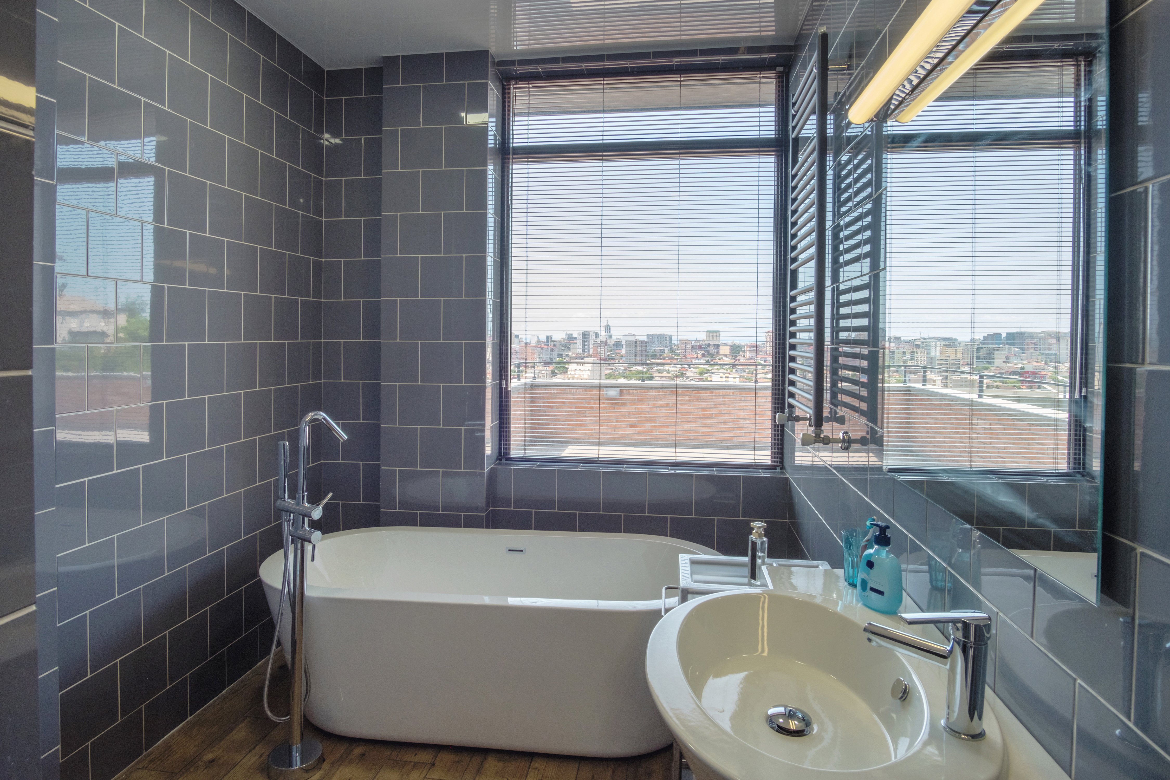 Gray tile bathroom with white tub and sink next to large window overlooking cityscape