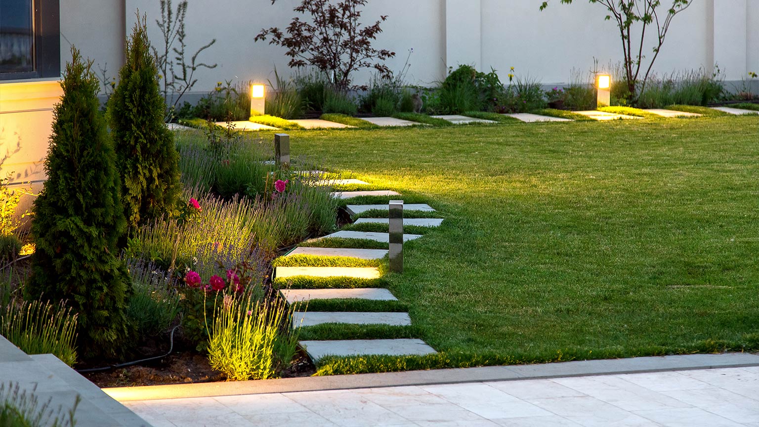 backyard with a flowerbed and green lawn with a marble walkway lined with garden lamp lighting