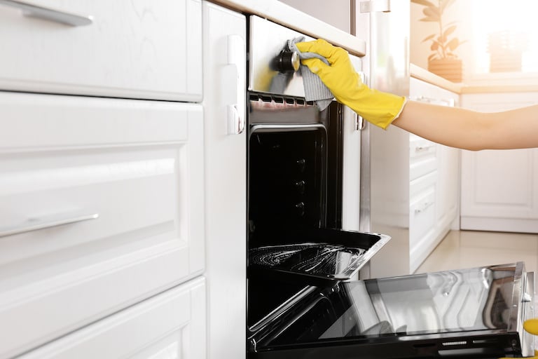 hand in yellow glove cleans black and stainless steel oven in kitchen with white cabinets