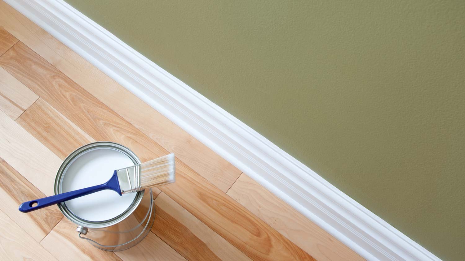 A paint bucket with a paintbrush on top of it next to a white baseboard