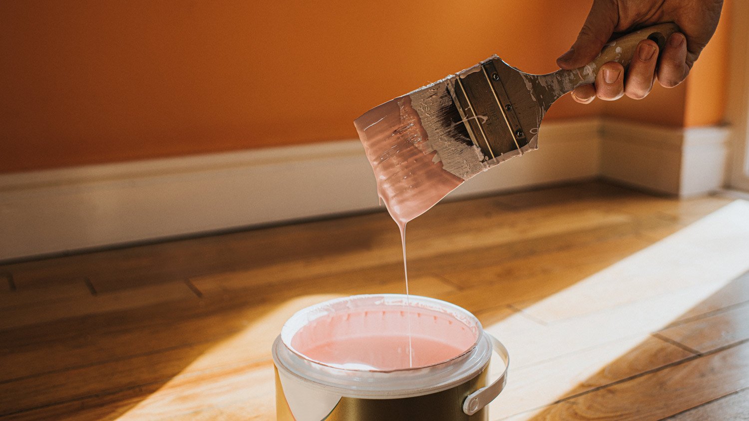 Hand dipping a paint brush into a large tin of pink paint as it drips back into the pot