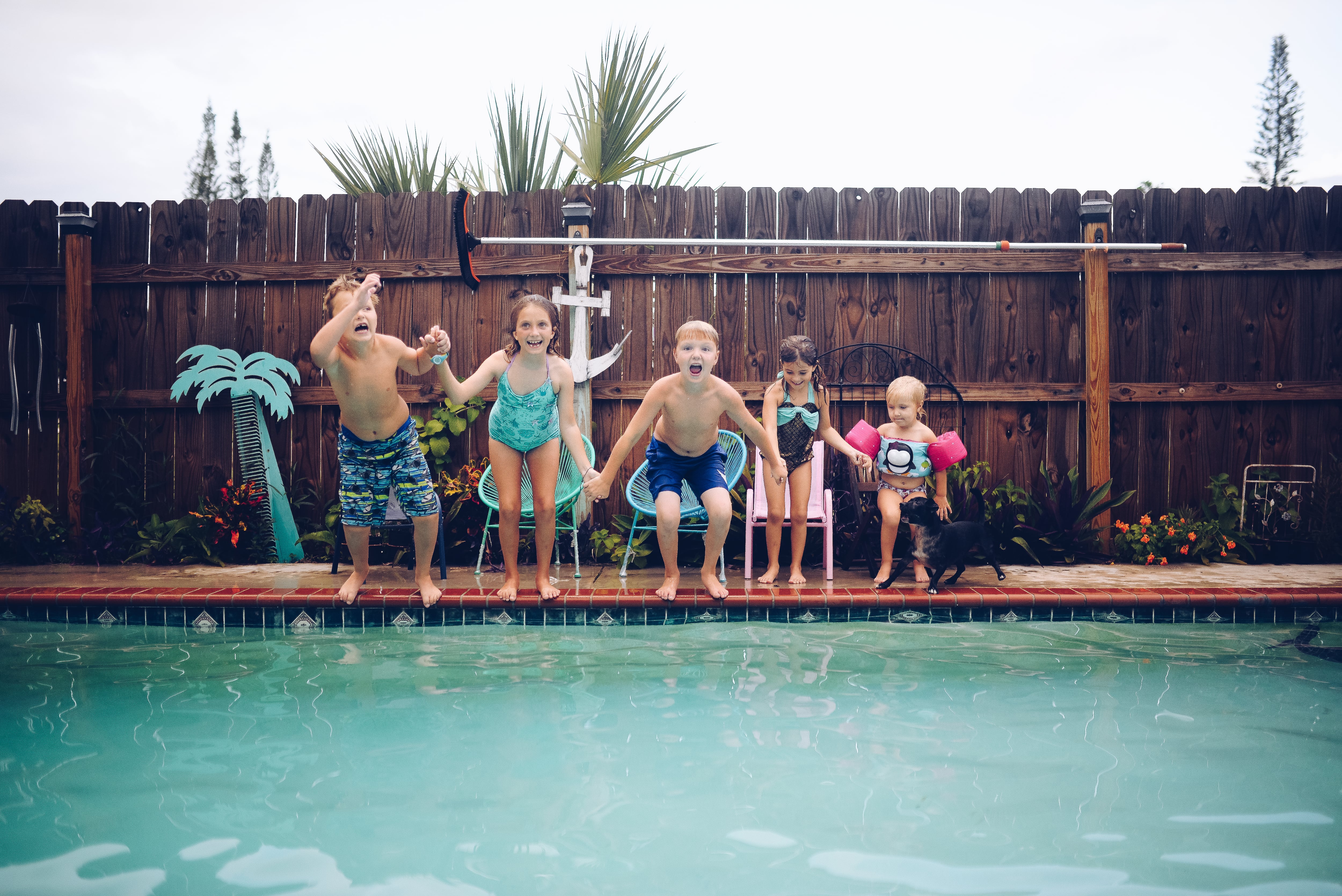 A group of children holding hands before jumping to a pool surrounded by a wooden fence