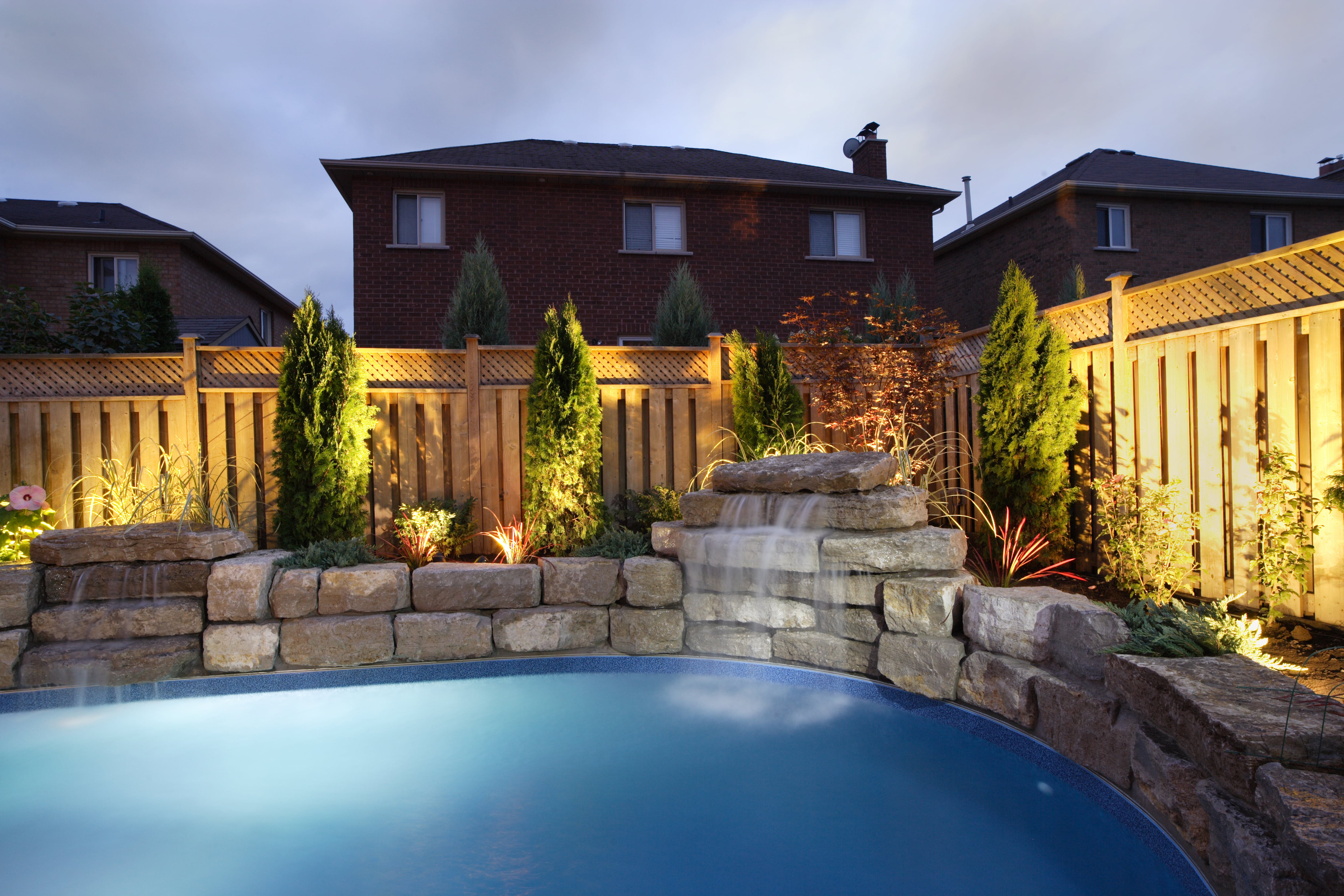 An inground pool at night with a small waterfall enclosed by a lattice top wooden fence