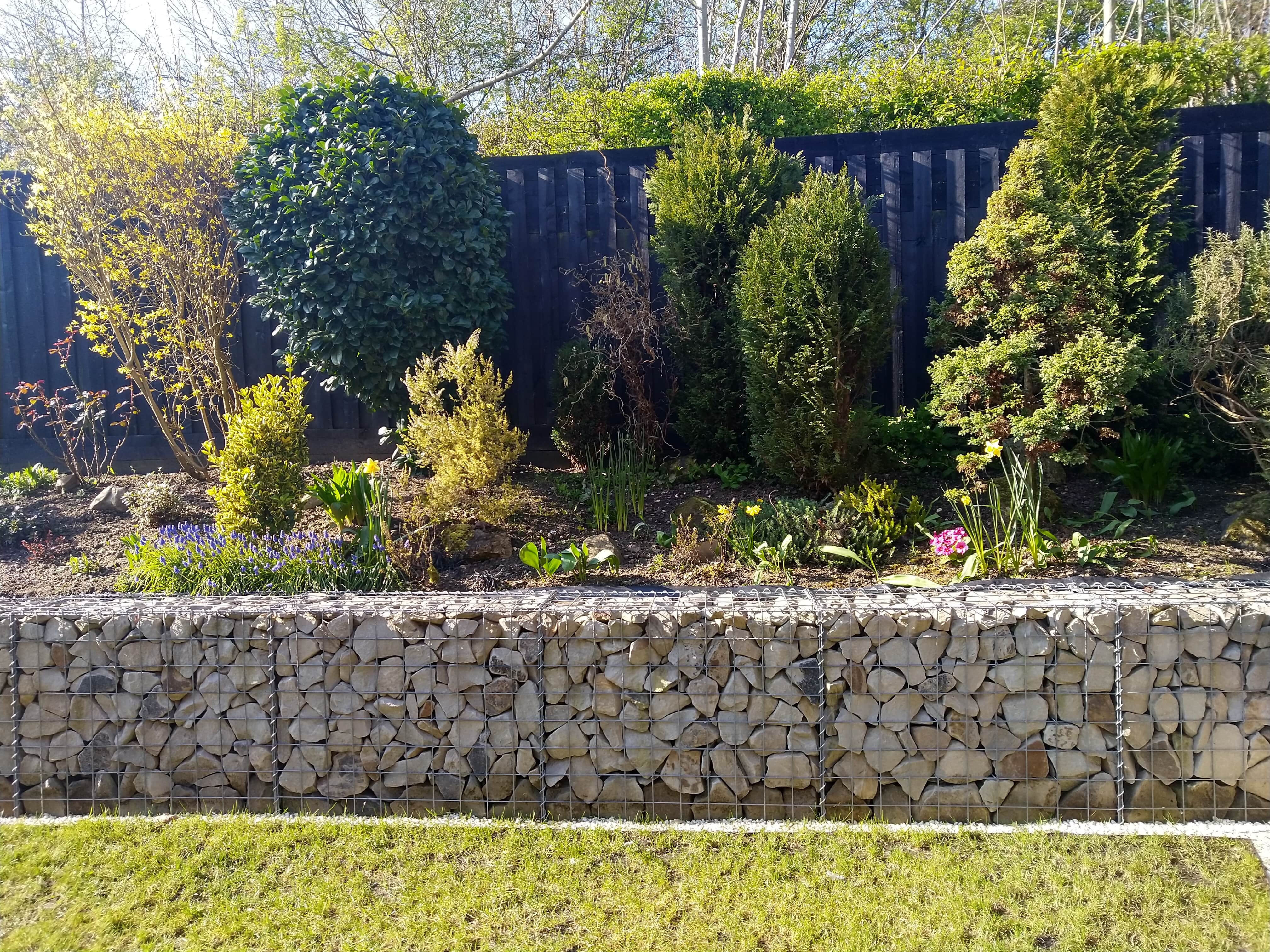 A gabion retaining wall in front of a large flower bed and steel fence