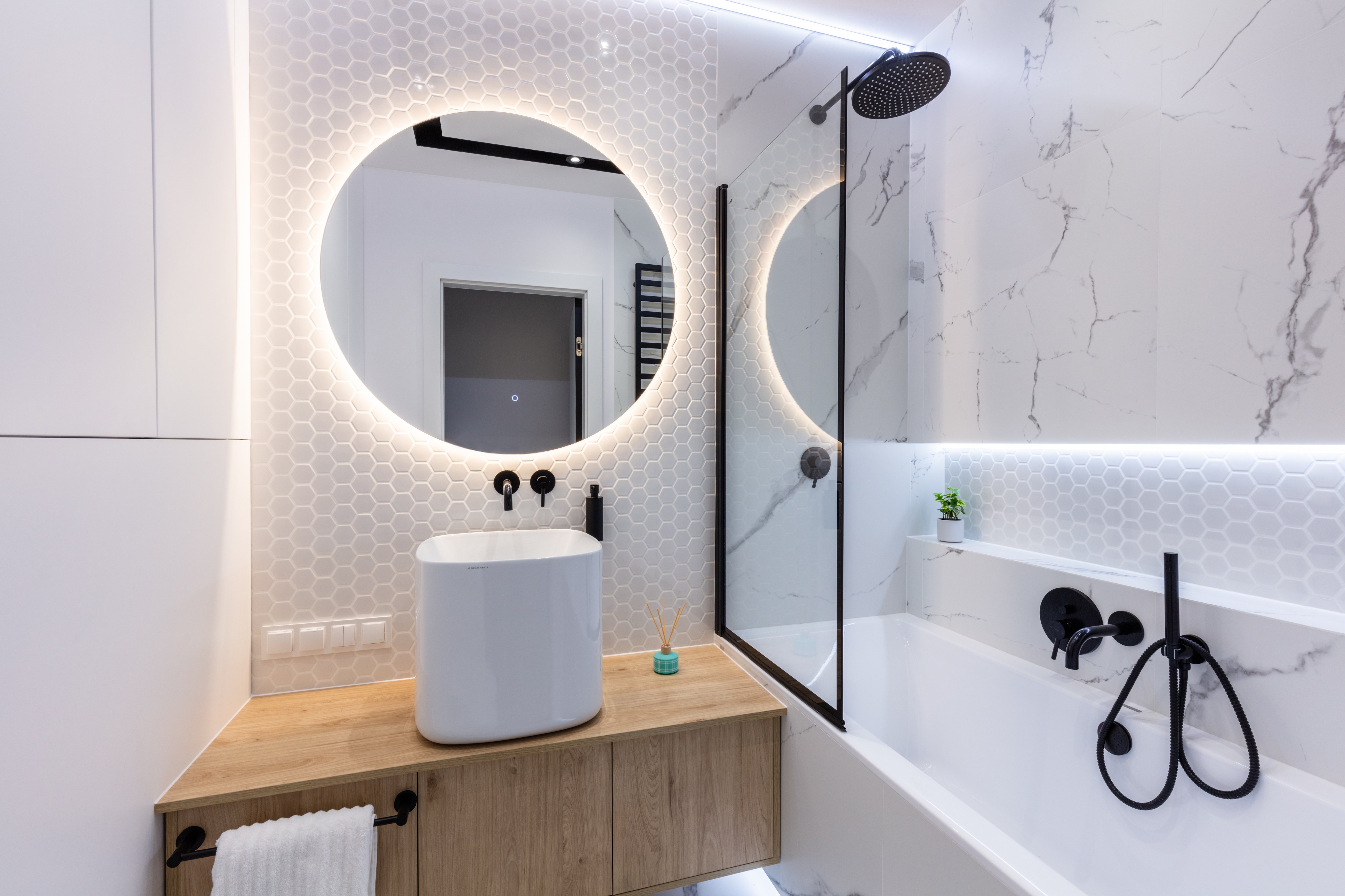 Modern small bathroom with backlit mirror and shower walls, marble tub and shower wall, wood sink, and black metal accents