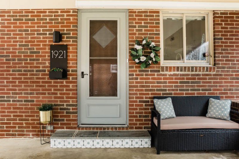 full entry way featuring gold plant stand, tiled step, patio bench, house number plant box and sconce light, and wreath (Photo by © Carrie Waller)