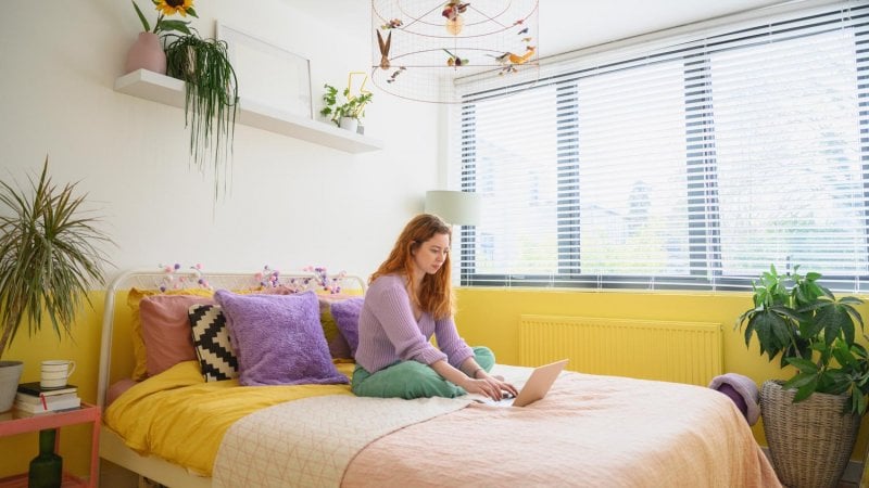 Woman relaxing in her yellow painted bedroom with her laptop (Photo by JohnnyGreig/E+ via Getty Images)