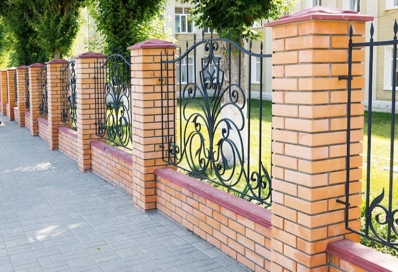 a tan brick fence with alternating metal grille design and stone sidewalk (Photo by olgavolodina - stock.adobe.com)
