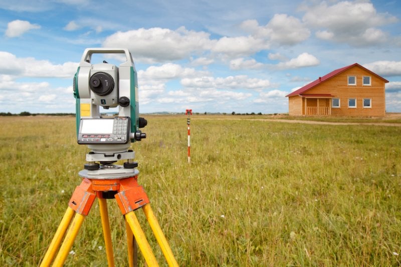 a metering device surveying land in a large field with a house in the distance (Photo by © Grispb - stock.adobe.com)