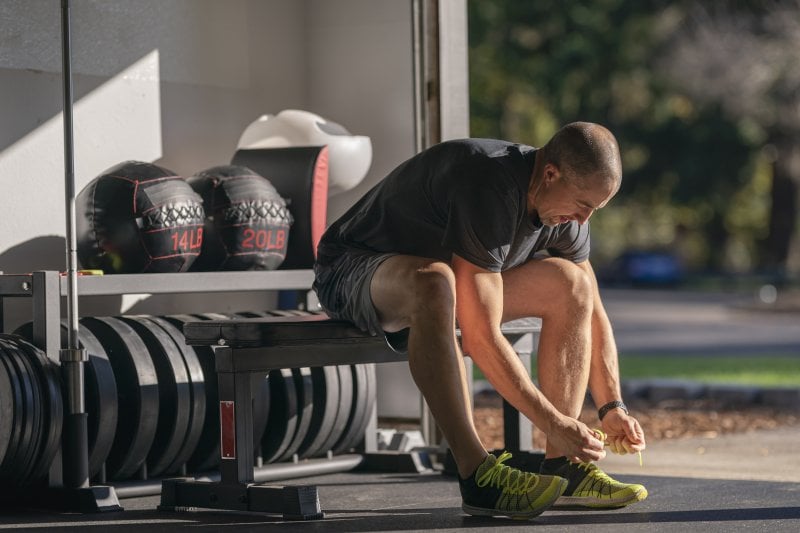 Man in gym clothes lacing up his sneakers in his garage home gym with gym equipment stored behind him (Photo by Fly View Productions / E+ via Getty Images)