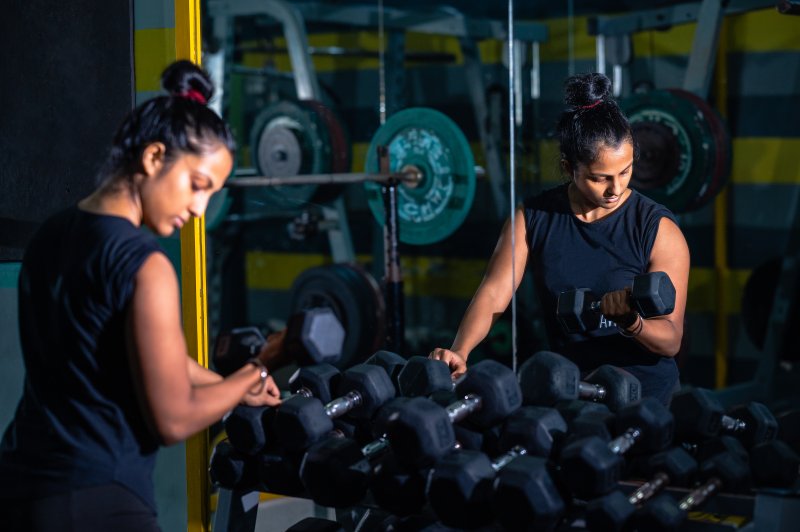 Woman leaning on weight rack with a dumbbell in her right hand doing a curl facing a large mirror in her garage home gym (Photo by Ankit Sah / E+ via Getty Images)