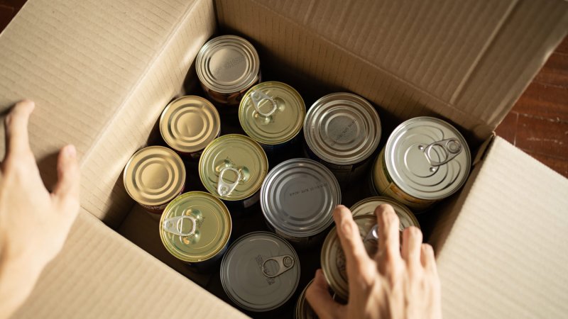 A hand placing canned food in a cardboard box (Photo by Wachiwit/iStock / Getty Images Plus via Getty Images)