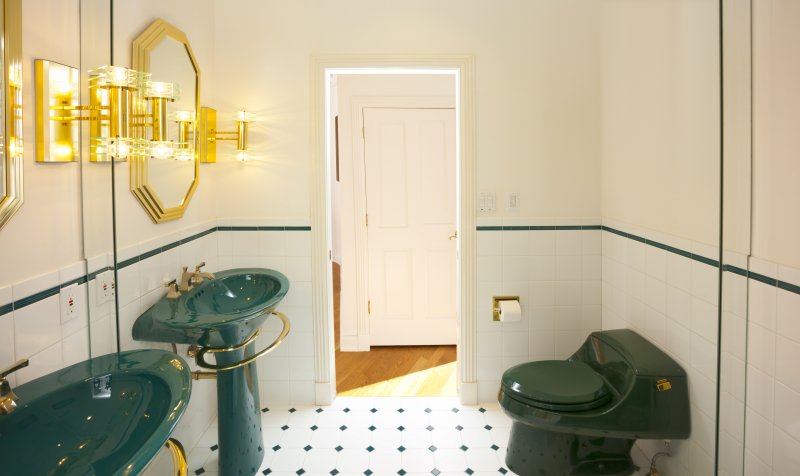Bathroom with green sink and toilet, gold metal accents, black-and-white tile floors, and a mirror covering the back wall (Photo by James Brey / iStock / Getty Images Plus via Getty Images)