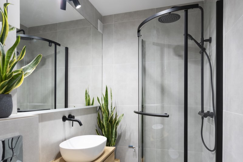 Small bathroom with gray tiles, glass and black metal shower, wall-mounted faucet, vessel sink, and plants (Photo by photosbysabkapl – stock.adobe.com)