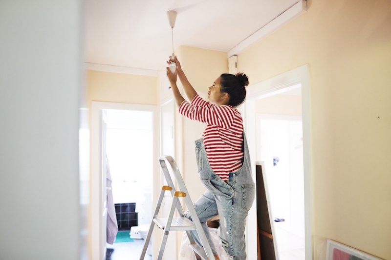 woman in overalls stands on a ladder and change hanging lightbulb from ceiling (Photo by Klaus Vedfelt/DigitalVision via Getty Images)