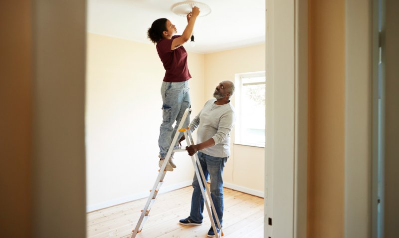 A woman installs a light bulb while her father holds the ladder (Photo by Klaus Vedfelt/DigitalVision via Getty Images)
